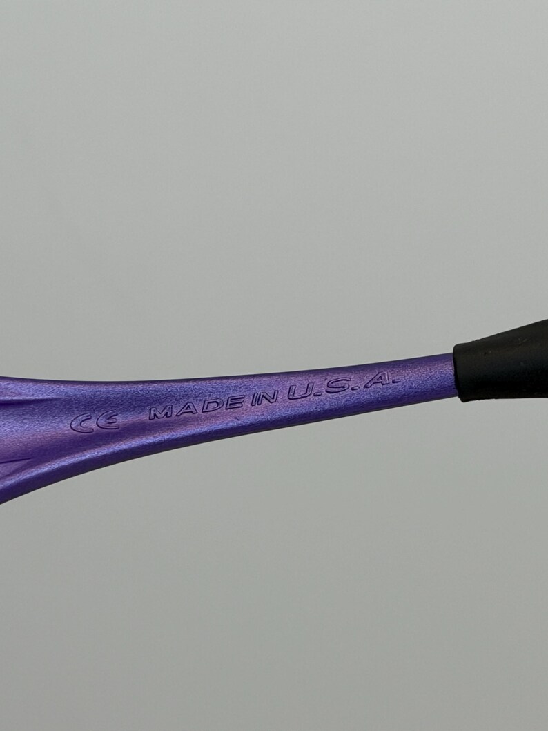 May include: Close-up of a purple eyeglass frame arm with the text "MADE IN U.S.A." and "CE" printed on it. The arm has a gradient color and a black tip.