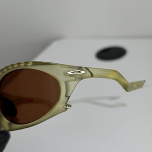 May include: Close-up of olive green sunglasses with brown lenses. The sunglasses have a curved design with a silver logo on the temple. The frame is a translucent, light green colour.