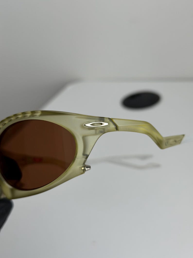 May include: Close-up of olive green sunglasses with brown lenses. The sunglasses have a curved design with a silver logo on the temple. The frame is a translucent, light green colour.