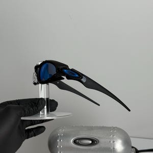 May include: Black and blue sports sunglasses with a unique, angular design. The sunglasses have blue lenses and are displayed on a clear stand. A silver case with the brand logo is also visible.