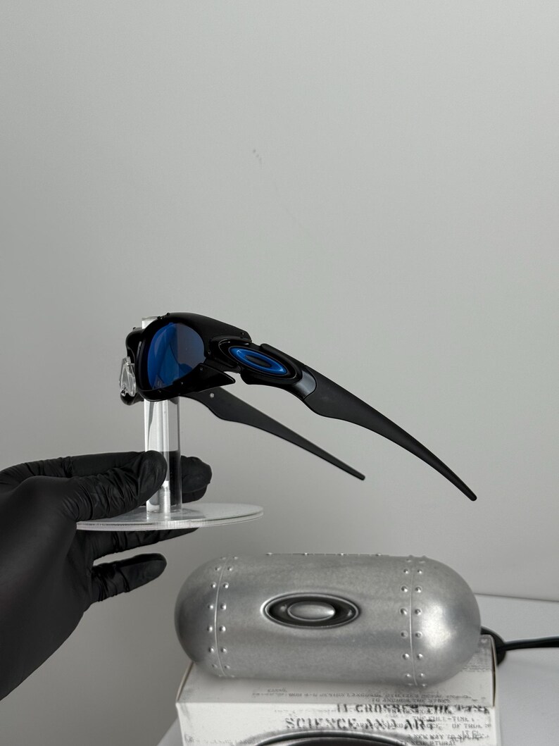 May include: Black and blue sports sunglasses with a unique, angular design. The sunglasses have blue lenses and are displayed on a clear stand. A silver case with the brand logo is also visible.