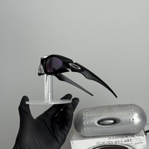 May include: Black sunglasses with a sleek, aerodynamic design, featuring dark lenses and a distinctive frame. The sunglasses are displayed on a clear stand, with a silver case and a box with the text "SCIENCE AND ART" visible in the background.
