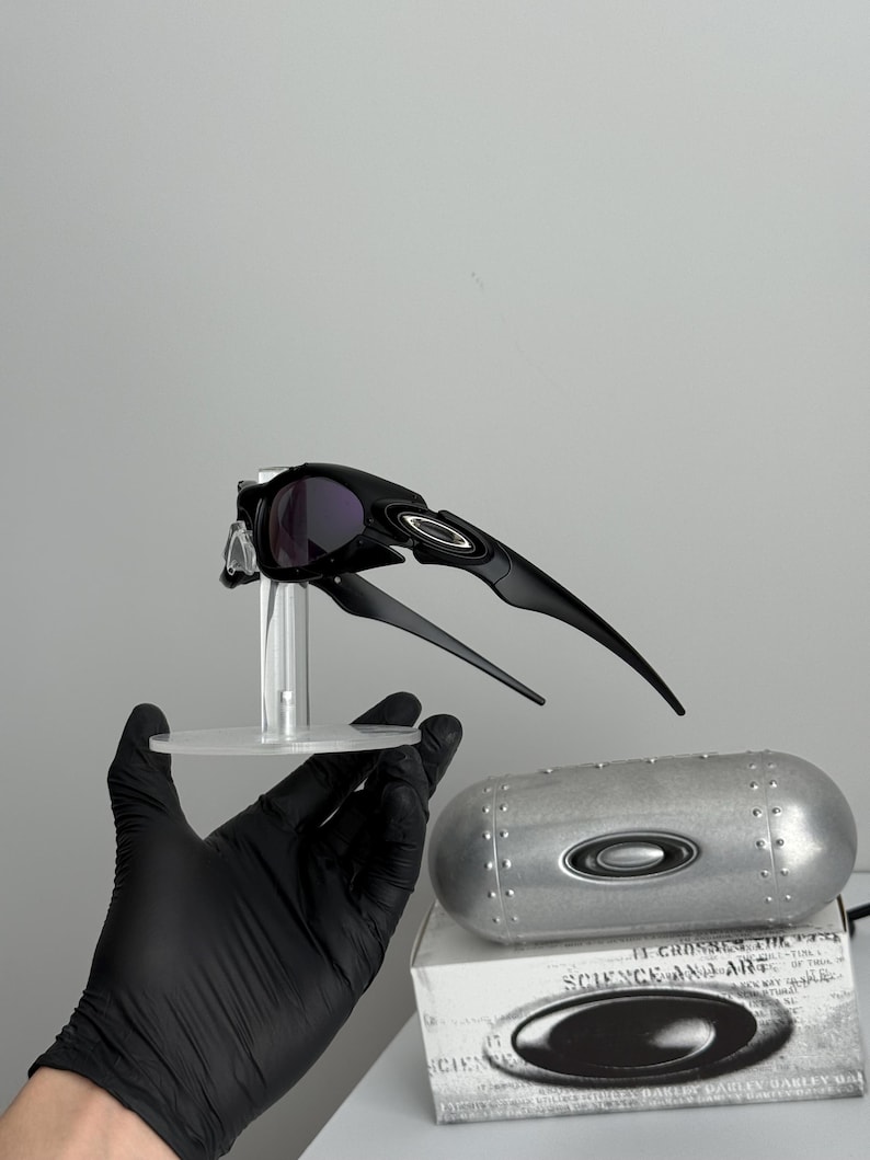 May include: Black sunglasses with a sleek, aerodynamic design, featuring dark lenses and a distinctive frame. The sunglasses are displayed on a clear stand, with a silver case and a box with the text "SCIENCE AND ART" visible in the background.