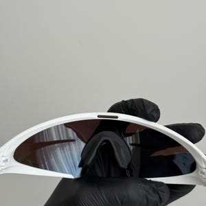 Puede incluir: Gafas de sol deportivas blancas con lentes oscuras. Las gafas tienen un dise&ntilde;o curvo y un puente nasal. Las lentes tienen un tinte degradado. Las gafas son sostenidas por una persona con guantes negros.
