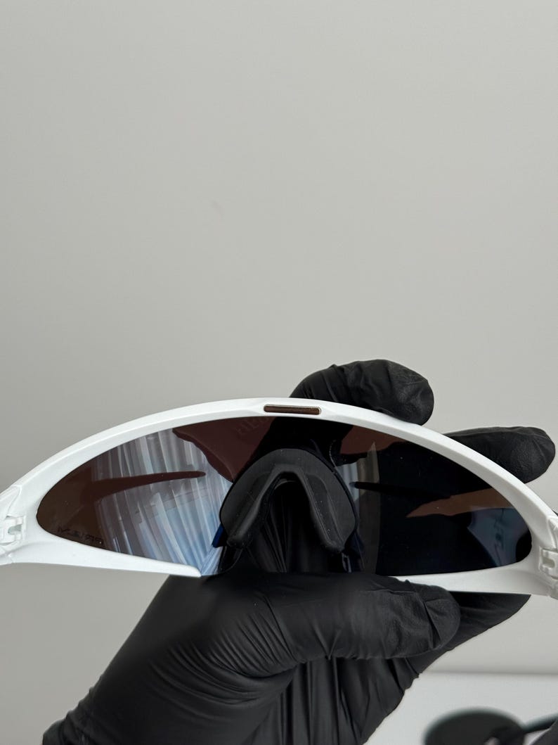 Puede incluir: Gafas de sol deportivas blancas con lentes oscuras. Las gafas tienen un dise&ntilde;o curvo y un puente nasal. Las lentes tienen un tinte degradado. Las gafas son sostenidas por una persona con guantes negros.