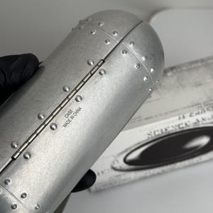 May include: Silver-colored, riveted case with a hinged design. The case has a cylindrical shape and features the text "CASE MADE IN CHINA". The case is likely for eyewear or similar items.