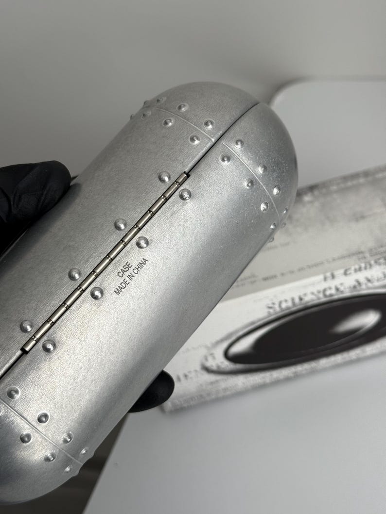 May include: Silver-colored, riveted case with a hinged design. The case has a cylindrical shape and features the text "CASE MADE IN CHINA". The case is likely for eyewear or similar items.