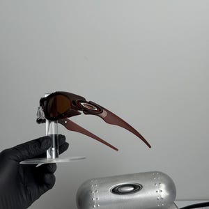 May include: Brown and black Oakley sunglasses with a unique, angular design. The sunglasses are displayed on a clear stand, with a silver case and a box visible in the background. The case has the Oakley logo.