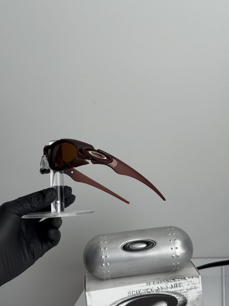 May include: Brown and black Oakley sunglasses with a unique, angular design. The sunglasses are displayed on a clear stand, with a silver case and a box visible in the background. The case has the Oakley logo.