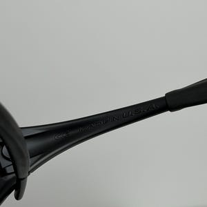 May include: Close-up of black sunglasses arm with the text "MADE IN U.S.A." printed in white. The sunglasses arm is made of a matte black material. The image is set against a plain, light background.