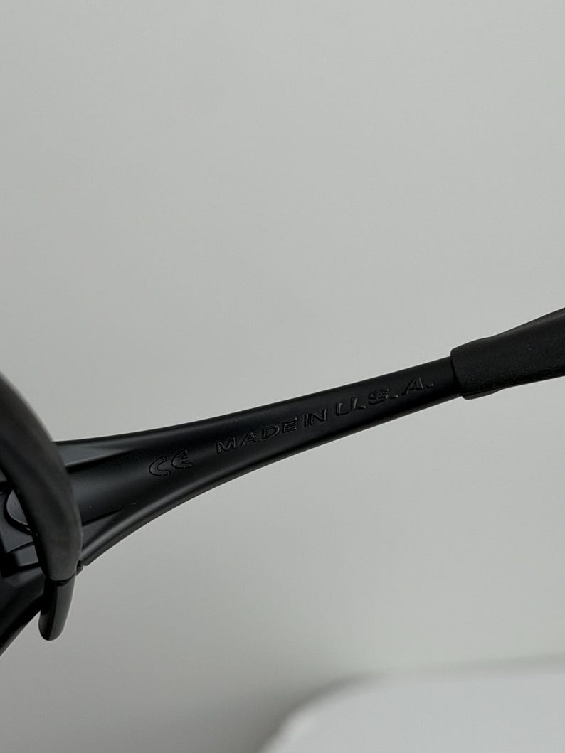 May include: Close-up of black sunglasses arm with the text "MADE IN U.S.A." printed in white. The sunglasses arm is made of a matte black material. The image is set against a plain, light background.