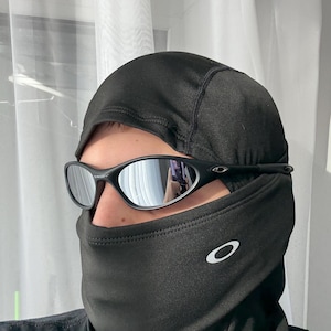 K&ouml;nnte beinhalten: Schwarze Sonnenbrille mit verspiegelten Gl&auml;sern, getragen &uuml;ber einer schwarzen Sturmhaube. Die Sturmhaube hat ein wei&szlig;es Logo auf der Vorderseite. Die Sonnenbrille hat einen schwarzen Rahmen.