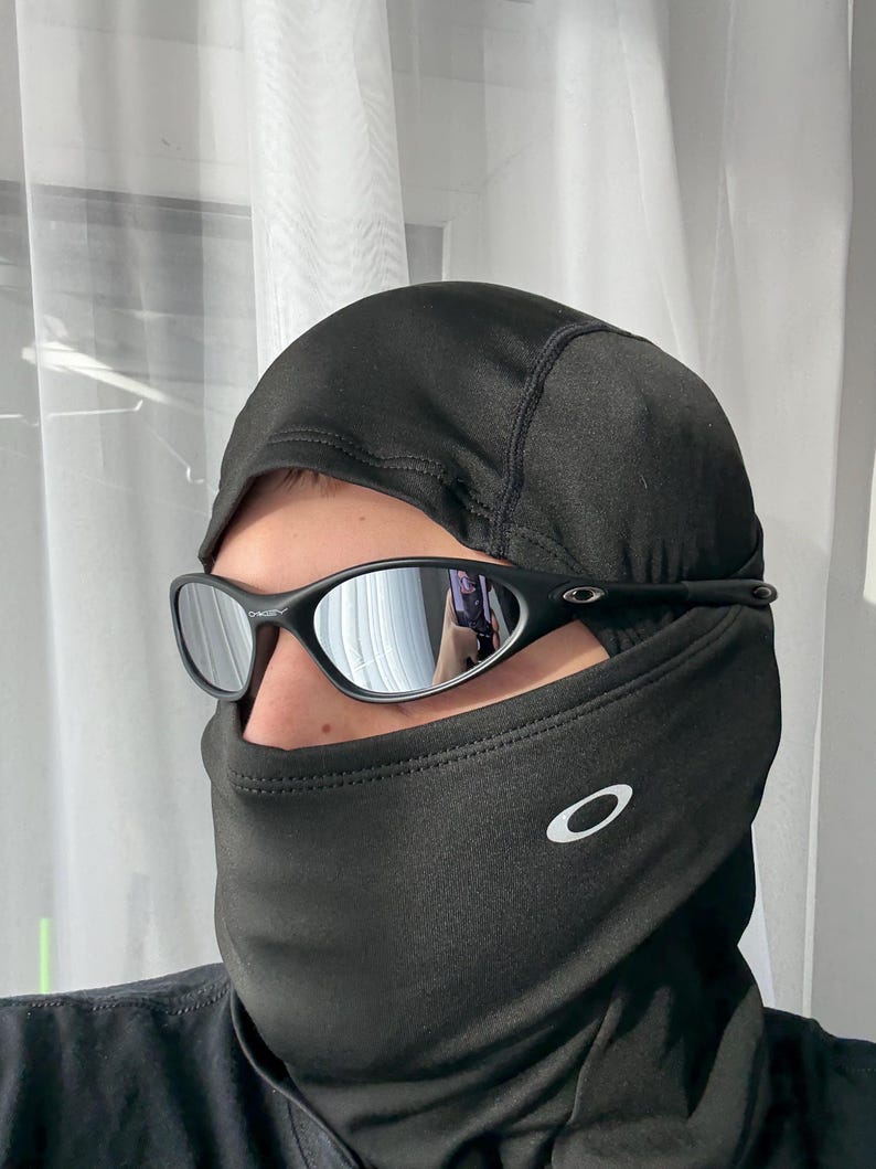 K&ouml;nnte beinhalten: Schwarze Sonnenbrille mit verspiegelten Gl&auml;sern, getragen &uuml;ber einer schwarzen Sturmhaube. Die Sturmhaube hat ein wei&szlig;es Logo auf der Vorderseite. Die Sonnenbrille hat einen schwarzen Rahmen.