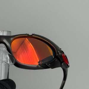 May include: Close-up of black sunglasses with red-tinted lenses. The sunglasses have a sleek, modern design with a curved frame and a small red accent on the side. The lenses reflect the surrounding environment.