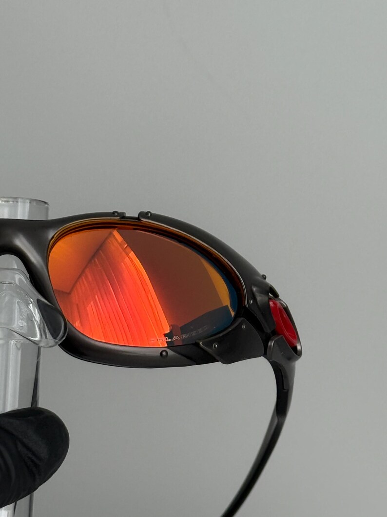 May include: Close-up of black sunglasses with red-tinted lenses. The sunglasses have a sleek, modern design with a curved frame and a small red accent on the side. The lenses reflect the surrounding environment.