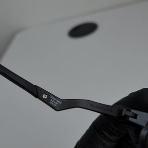 May include: Close-up of a black eyeglass frame. The frame has the text "PLANTARIS" and the numbers "09437-0463 63019 126 3P" printed on the arm. The frame is held by a gloved hand.