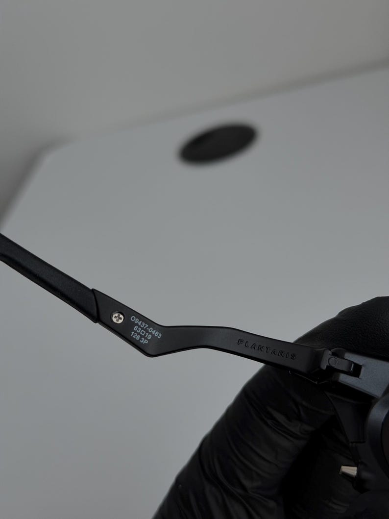 May include: Close-up of a black eyeglass frame. The frame has the text "PLANTARIS" and the numbers "09437-0463 63019 126 3P" printed on the arm. The frame is held by a gloved hand.