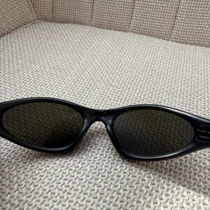 May include: Black sunglasses with dark lenses. The sunglasses have an oval shape and a matte finish. The arms of the sunglasses are thick and feature a subtle design. The sunglasses are resting on a textured, neutral-coloured surface.