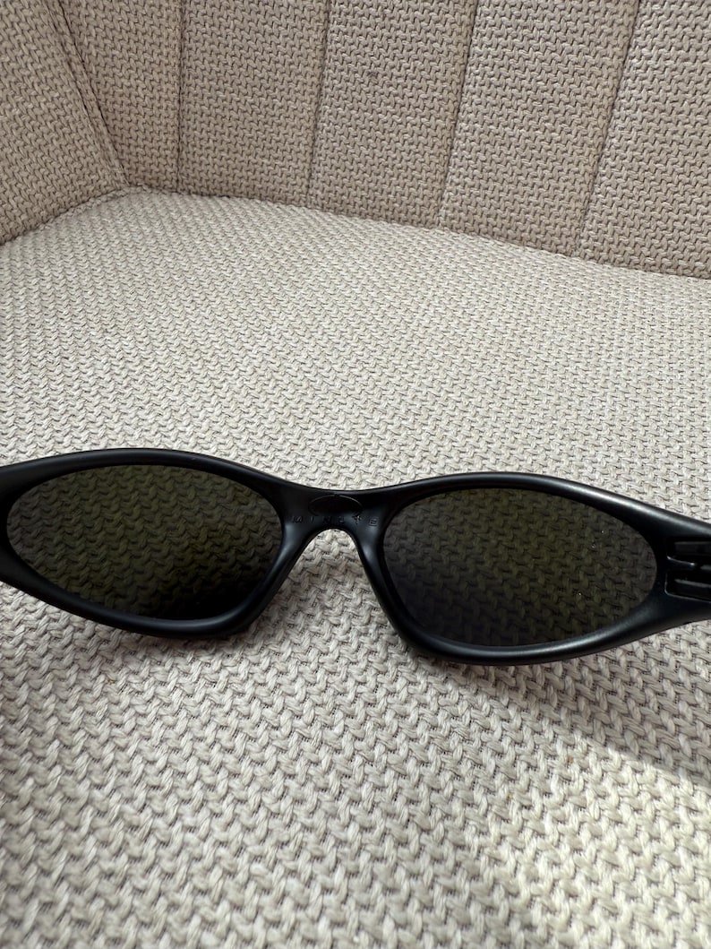 May include: Black sunglasses with dark lenses. The sunglasses have an oval shape and a matte finish. The arms of the sunglasses are thick and feature a subtle design. The sunglasses are resting on a textured, neutral-coloured surface.