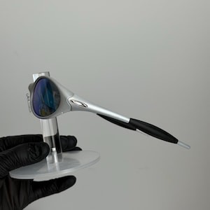 May include: Silver sunglasses with blue lenses. The sunglasses have a modern design with black earpieces. The glasses are displayed on a clear, circular stand.