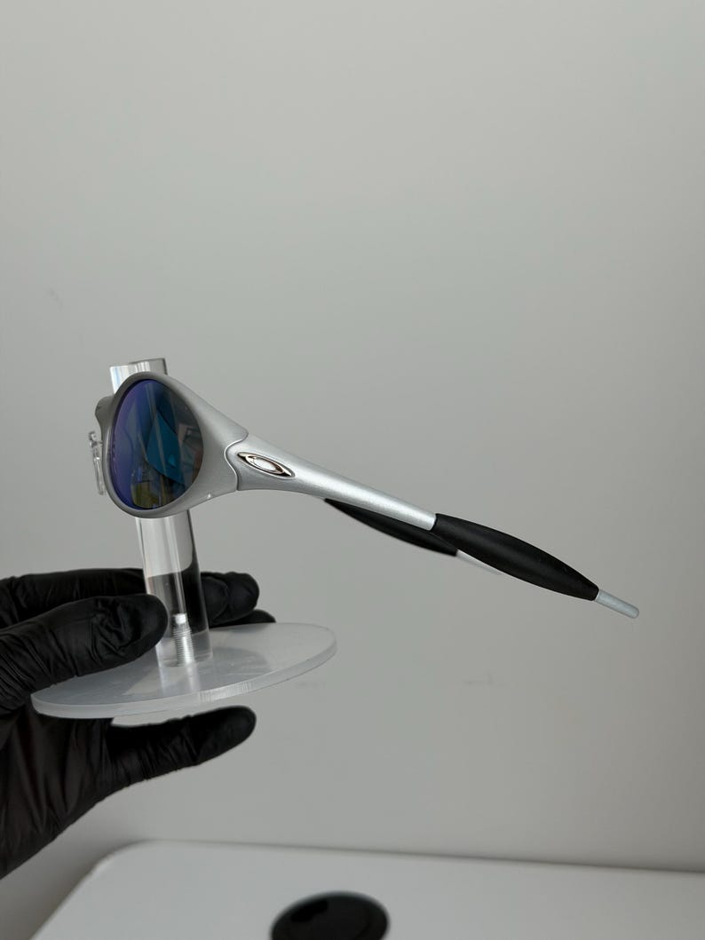 May include: Silver sunglasses with blue lenses. The sunglasses have a modern design with black earpieces. The glasses are displayed on a clear, circular stand.