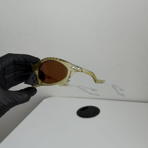 May include: Olive green sunglasses with brown lenses and white arms. The sunglasses have a sporty design with a curved frame and a small logo on the side. The lenses are a warm brown colour, and the arms are white.