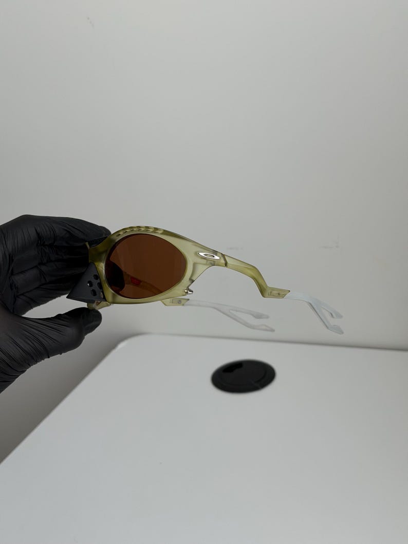 May include: Olive green sunglasses with brown lenses and white arms. The sunglasses have a sporty design with a curved frame and a small logo on the side. The lenses are a warm brown colour, and the arms are white.