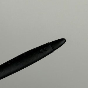 May include: Close-up of a black stylus with a rounded tip. The stylus has a matte finish and a subtle CE marking near the tip. The background is a solid, neutral gray.