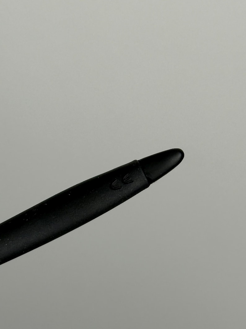 May include: Close-up of a black stylus with a rounded tip. The stylus has a matte finish and a subtle CE marking near the tip. The background is a solid, neutral gray.