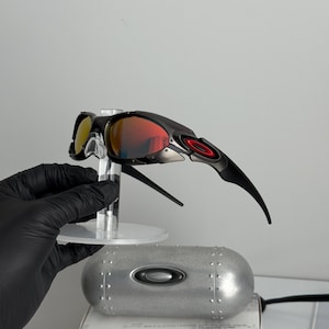 May include: Black and red sunglasses with a mirrored lens. The sunglasses have a sleek, modern design with a black frame and red accents. The lenses are a vibrant red color. The sunglasses are displayed on a clear stand.