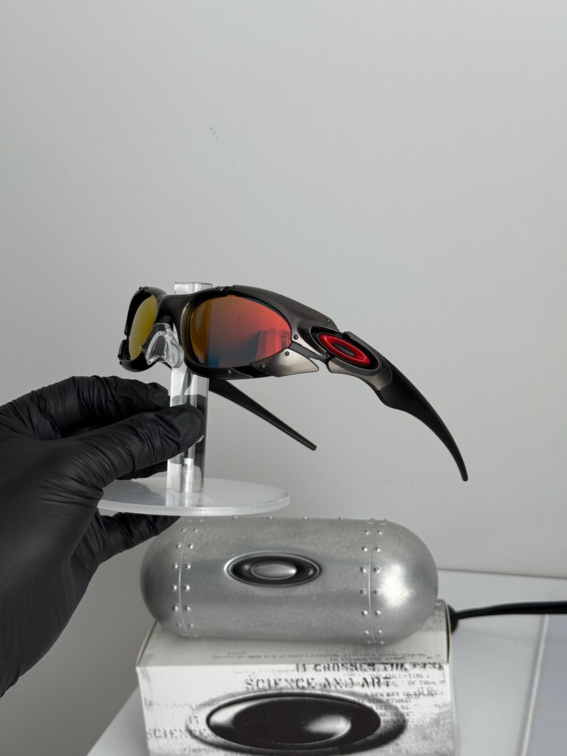 May include: Black and red sunglasses with a mirrored lens. The sunglasses have a sleek, modern design with a black frame and red accents. The lenses are a vibrant red color. The sunglasses are displayed on a clear stand.