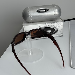 May include: Brown-framed sunglasses with brown lenses are displayed on a clear stand. A silver, cylindrical case with the Oakley logo is behind the sunglasses. A box with the text "SCIENCE AND ART" is also visible.