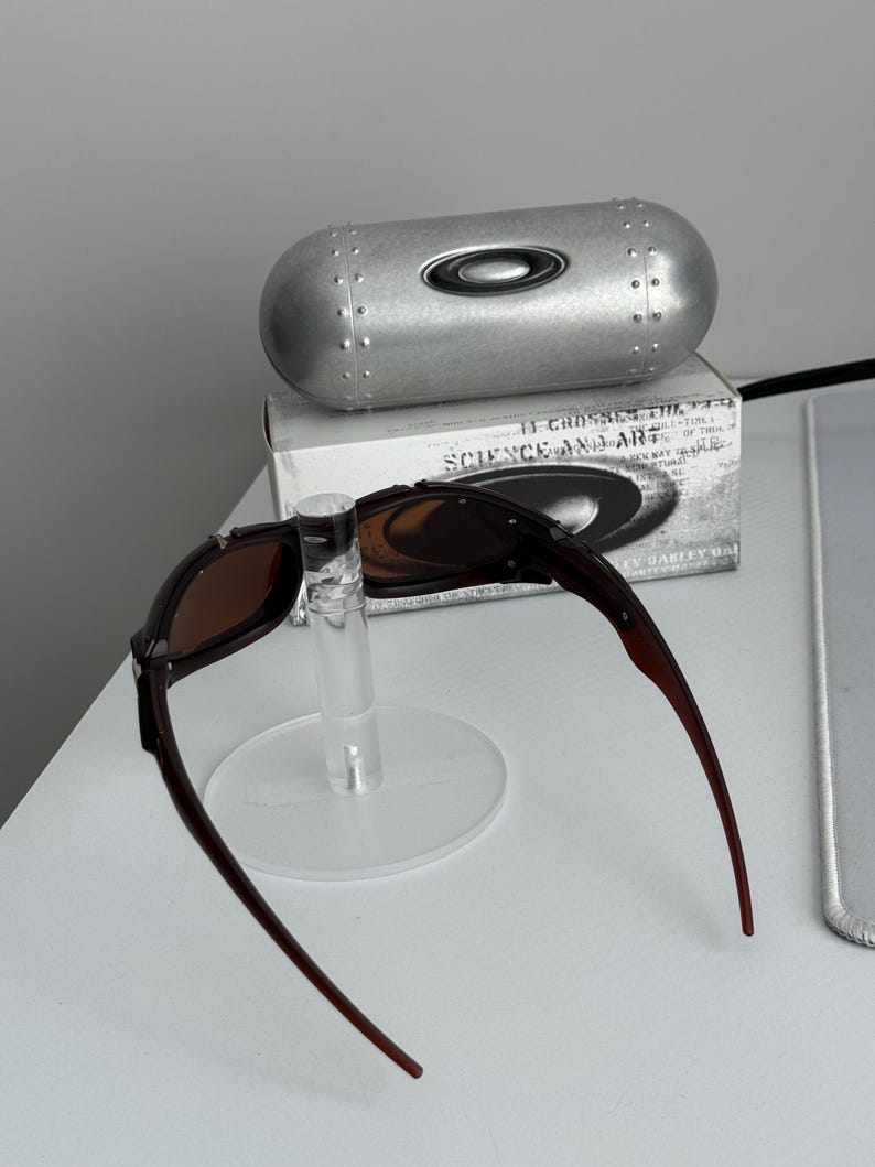 May include: Brown-framed sunglasses with brown lenses are displayed on a clear stand. A silver, cylindrical case with the Oakley logo is behind the sunglasses. A box with the text "SCIENCE AND ART" is also visible.
