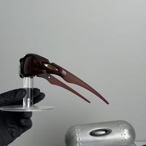 May include: Brown sunglasses with a unique, angular design, held on a clear stand. The sunglasses have a sleek, modern aesthetic. A silver case and a box with the text "SCIENCE AND ART" are also visible.