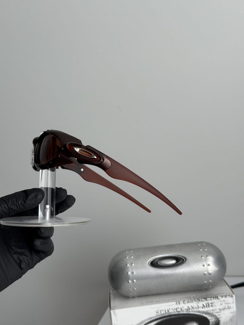 May include: Brown sunglasses with a unique, angular design, held on a clear stand. The sunglasses have a sleek, modern aesthetic. A silver case and a box with the text "SCIENCE AND ART" are also visible.