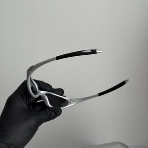 Puede incluir: Gafas de sol con montura plateada, lentes negras y puntas de patillas negras. Las gafas son sujetadas por una mano enguantada, con un estuche plateado en el fondo.