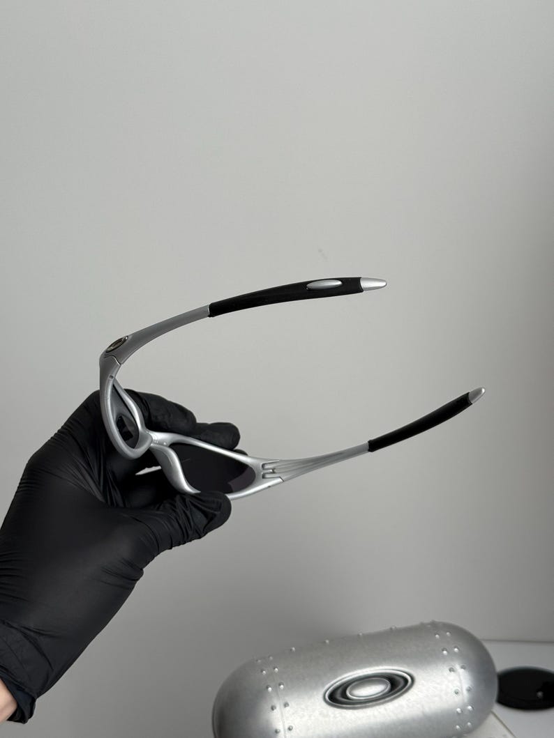 Puede incluir: Gafas de sol con montura plateada, lentes negras y puntas de patillas negras. Las gafas son sujetadas por una mano enguantada, con un estuche plateado en el fondo.