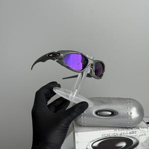 May include: Gray and black sunglasses with purple lenses are displayed on a clear stand. The sunglasses have a sleek, modern design. A silver case and a white box with the words "SCIENCE AND ART" are also visible.