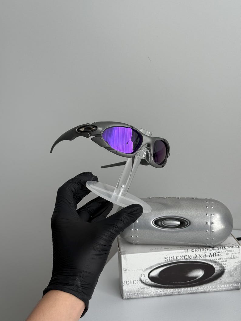 May include: Gray and black sunglasses with purple lenses are displayed on a clear stand. The sunglasses have a sleek, modern design. A silver case and a white box with the words "SCIENCE AND ART" are also visible.