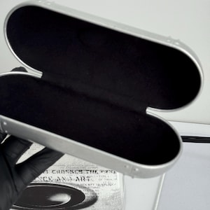 May include: Open silver eyeglasses case with a black interior. The case is oval-shaped and has a hinged design. The case is empty and ready to hold a pair of glasses. The case is sitting on top of a box with text.