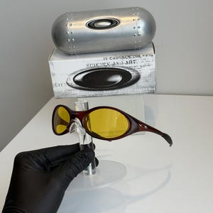 K&ouml;nnte beinhalten: Braune und gelb get&ouml;nte Oakley-Sonnenbrille mit passendem Etui und Box. Die Sonnenbrille hat einen geschwungenen Rahmen und ein schlankes Design. Das Etui ist silberfarben und zylindrisch. Auf der Box steht "SCIENCE AND ART".