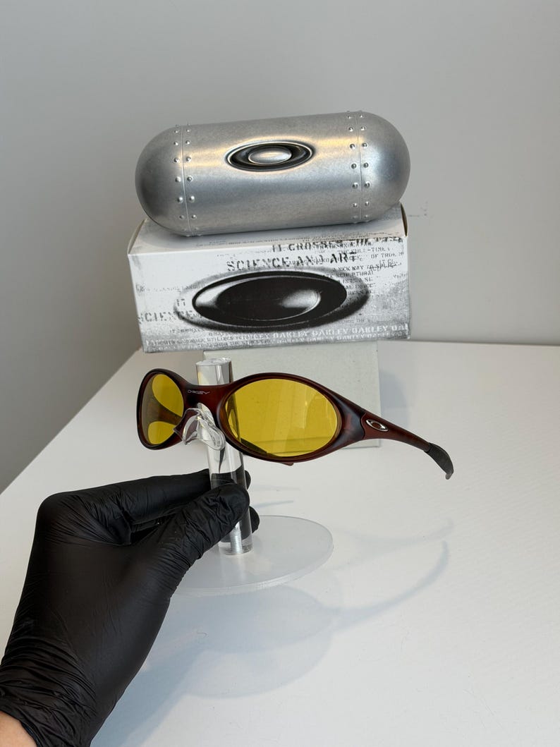K&ouml;nnte beinhalten: Braune und gelb get&ouml;nte Oakley-Sonnenbrille mit passendem Etui und Box. Die Sonnenbrille hat einen geschwungenen Rahmen und ein schlankes Design. Das Etui ist silberfarben und zylindrisch. Auf der Box steht "SCIENCE AND ART".