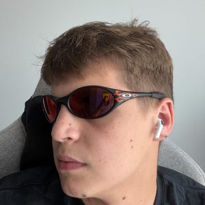May include: Black oval sunglasses with red lenses and flame accents on the temples. The sunglasses have a sleek, sporty design. The person is wearing a black t-shirt and white earbuds.
