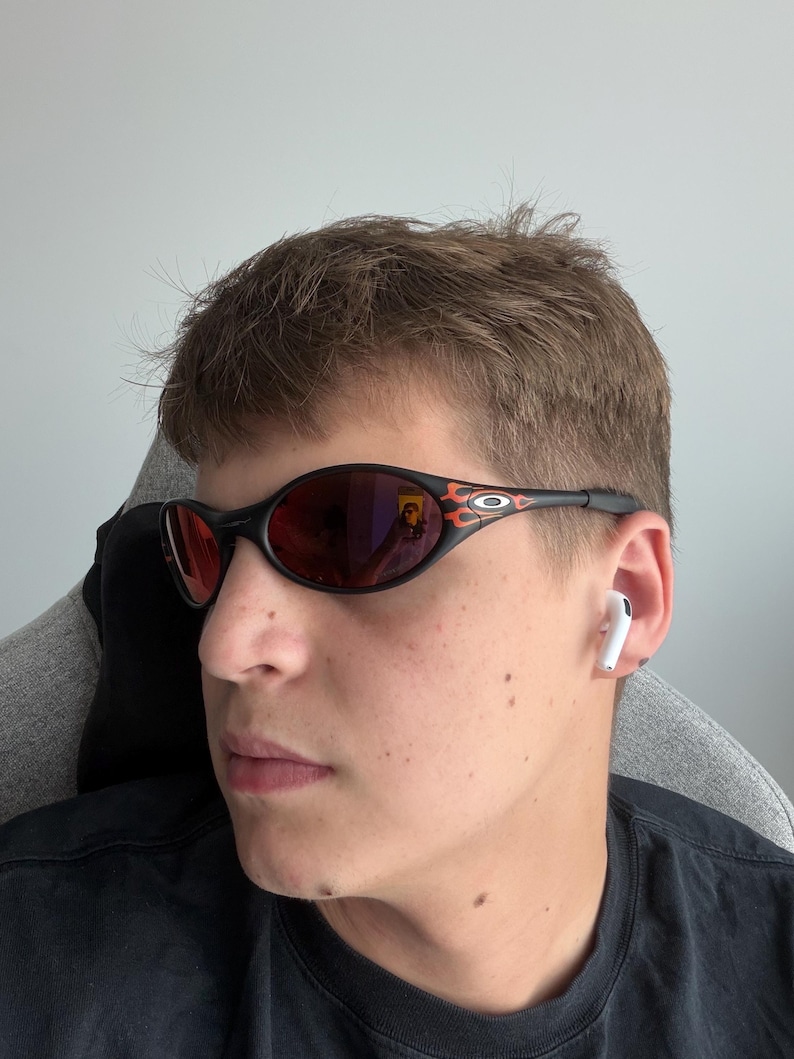May include: Black oval sunglasses with red lenses and flame accents on the temples. The sunglasses have a sleek, sporty design. The person is wearing a black t-shirt and white earbuds.