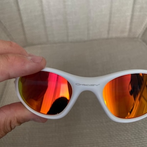 Puede incluir: Gafas de sol blancas con lentes espejadas rojo-naranja. Las gafas tienen una montura curvada y la marca "OAKLEY" est&aacute; impresa en la montura. Las lentes reflejan el entorno.
