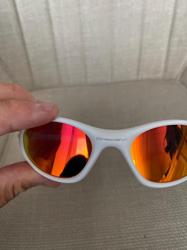 Puede incluir: Gafas de sol blancas con lentes espejadas rojo-naranja. Las gafas tienen una montura curvada y la marca "OAKLEY" est&aacute; impresa en la montura. Las lentes reflejan el entorno.
