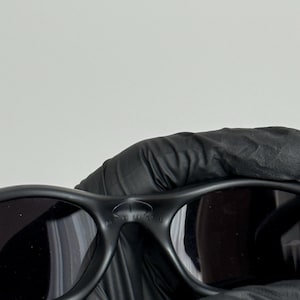 K&ouml;nnte beinhalten: Schwarze Sonnenbrille mit dunklen Gl&auml;sern. Die Sonnenbrille hat eine matte Oberfl&auml;che und einen geschwungenen Rahmen. Das Wort "MUTE" ist auf dem Steg der Sonnenbrille zu sehen. Die Sonnenbrille wird von einer behandschuhten Hand gehalten.
