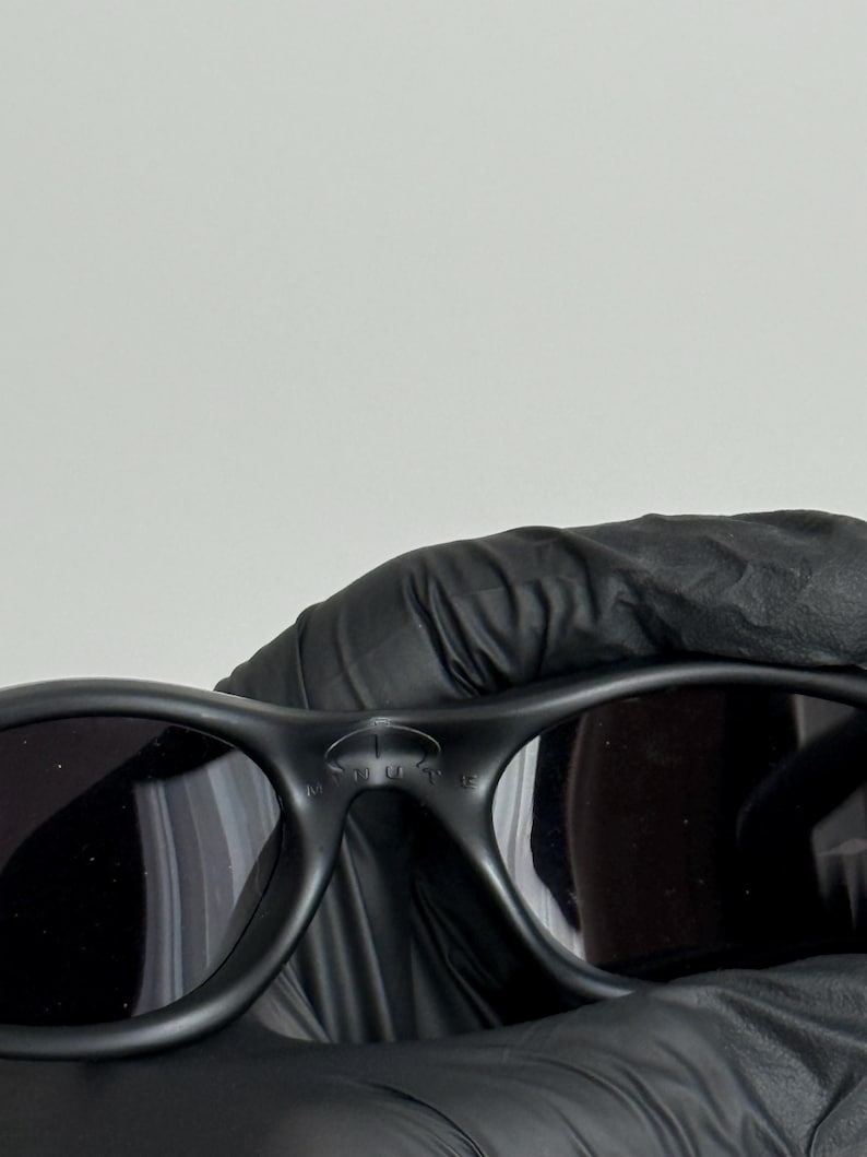 K&ouml;nnte beinhalten: Schwarze Sonnenbrille mit dunklen Gl&auml;sern. Die Sonnenbrille hat eine matte Oberfl&auml;che und einen geschwungenen Rahmen. Das Wort "MUTE" ist auf dem Steg der Sonnenbrille zu sehen. Die Sonnenbrille wird von einer behandschuhten Hand gehalten.