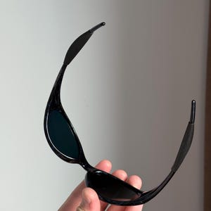 Puede incluir: Gafas de sol negras con lentes oscuras. Las gafas tienen una montura elegante y curvada, con patillas flexibles. Las lentes son oscuras y la montura parece brillante. Las patillas tienen una secci&oacute;n de goma para un ajuste seguro.