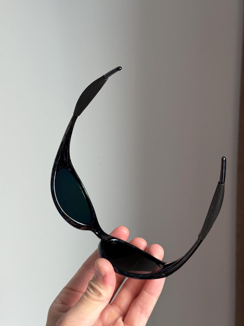Puede incluir: Gafas de sol negras con lentes oscuras. Las gafas tienen una montura elegante y curvada, con patillas flexibles. Las lentes son oscuras y la montura parece brillante. Las patillas tienen una secci&oacute;n de goma para un ajuste seguro.